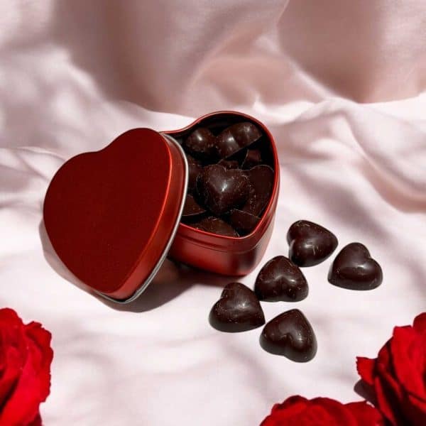 Ett rött plåthjärta fyllt med hjärtformade praliner av mörk choklad, liggande på en vit silkesslöja med röda rosor i bakgrunden.